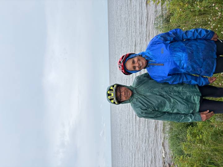 Couple in rain gear and helmets stands at a windy Baltic seashore under grey skies.