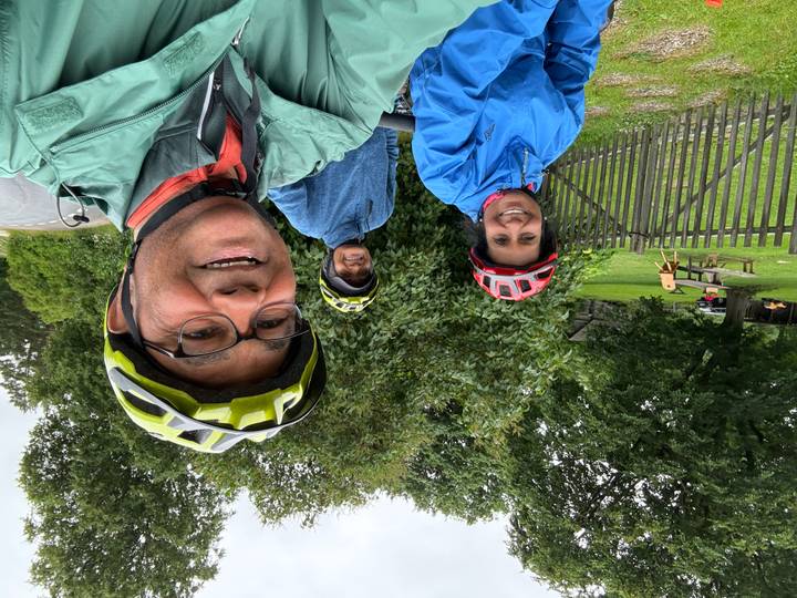 Cyclists smile for a selfie beside lush greenery after a ride on a rainy day.