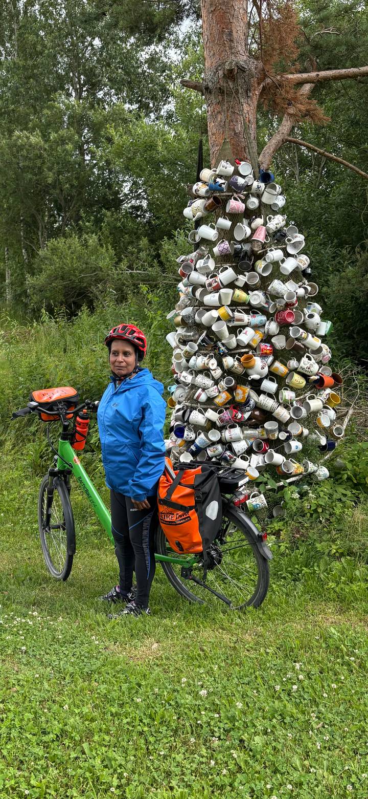 Cyclist in a blue rain jacket and red helmet stands with a green touring bike beside a sculptural tower of stacked coffee mugs in a lush, green setting.