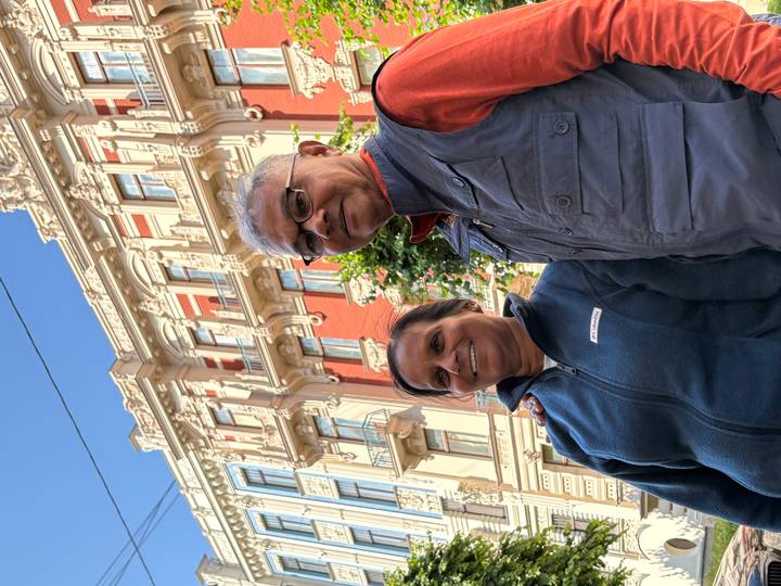Smiling couple poses in front of an ornate red-and-cream Art Nouveau façade under a clear blue sky.