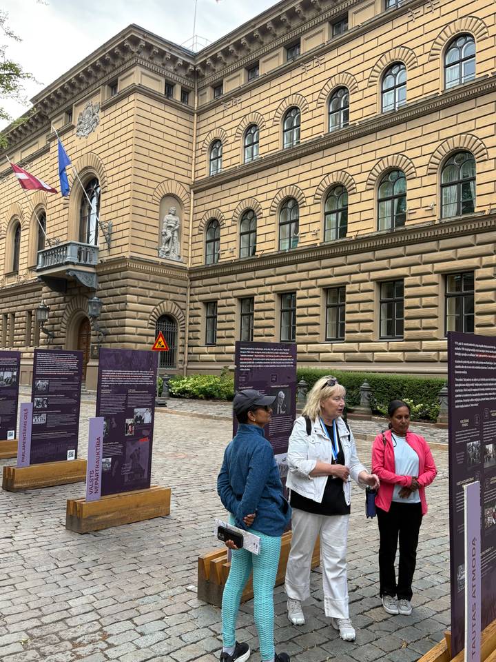 Small tour group listens to a guide outside a historic stone building with informational panels on a cobblestone street.