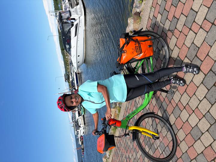 Cyclist with a red helmet and turquoise jersey poses with a touring bike on a harbour promenade lined with yachts under a bright blue sky.