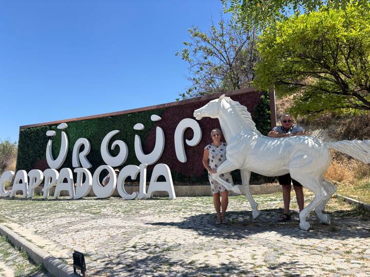 Two travelers pose beside a white horse statue by the Ürgüp Cappadocia sign on a sunny day.