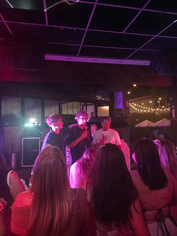 Young men singing on a small nightclub stage under purple and blue lighting while a crowd watches