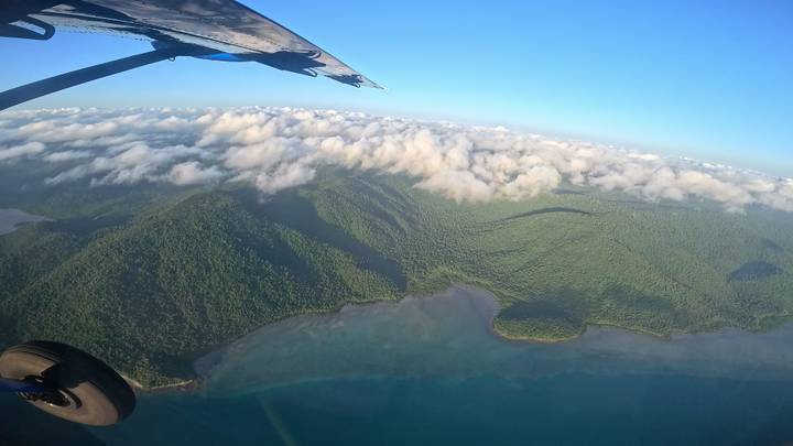 Aerial view from a small aircraft showing green rainforest covered mountains meeting turquoise coastal waters under scattered clouds