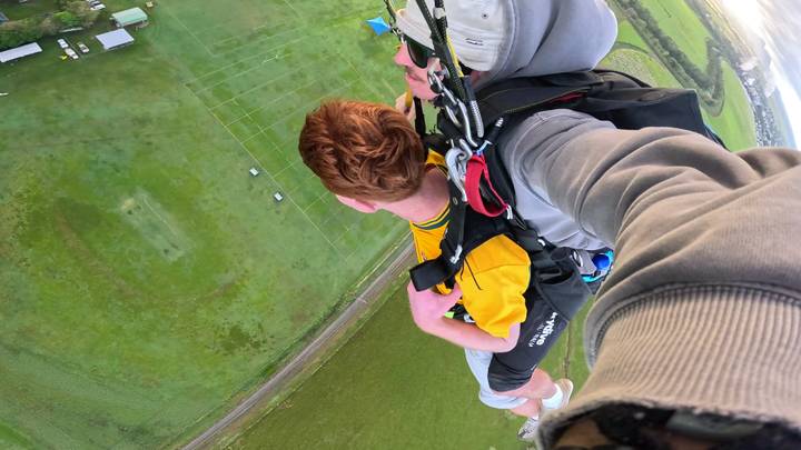 Tandem skydivers descend over green fields and farmland, captured from the harness perspective high above the ground