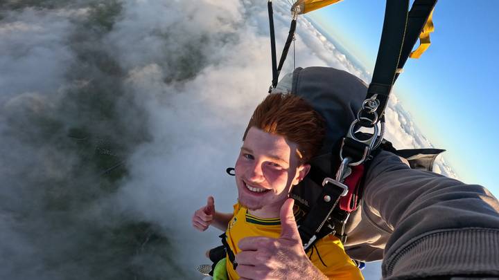 Smiling skydiver giving thumbs-up mid-air among white clouds with parachute straps visible