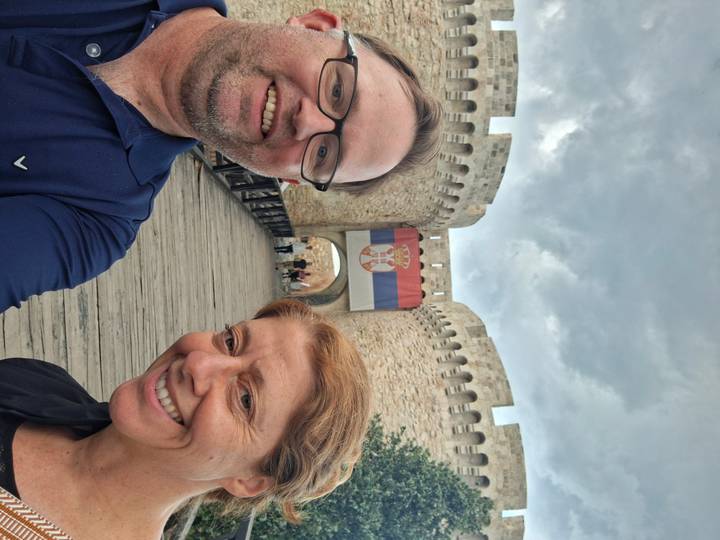 Selfie of smiling couple in front of Belgrade Fortress gate draped with the Serbian flag under cloudy skies.