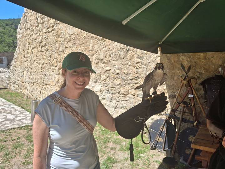 Smiling woman wearing a cap holds a falcon on a gloved hand beside a stone fortress wall and a small display stand.