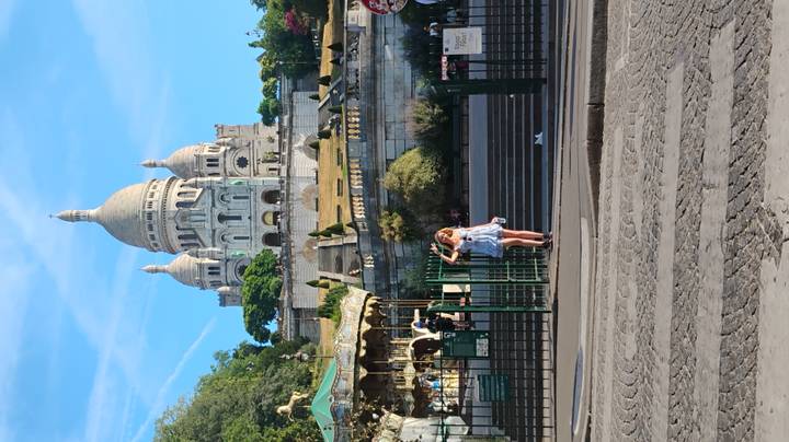 Woman poses at the base of Sacré-Cœur Basilica on Montmartre with carousel and gardens below.