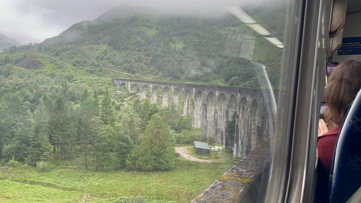 View from a train window of the long stone Glenfinnan Viaduct curving through a lush green valley.