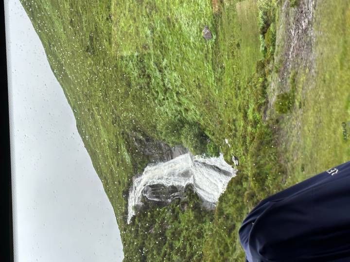 Small waterfall cascading down a green hillside viewed through a rain-spattered window.