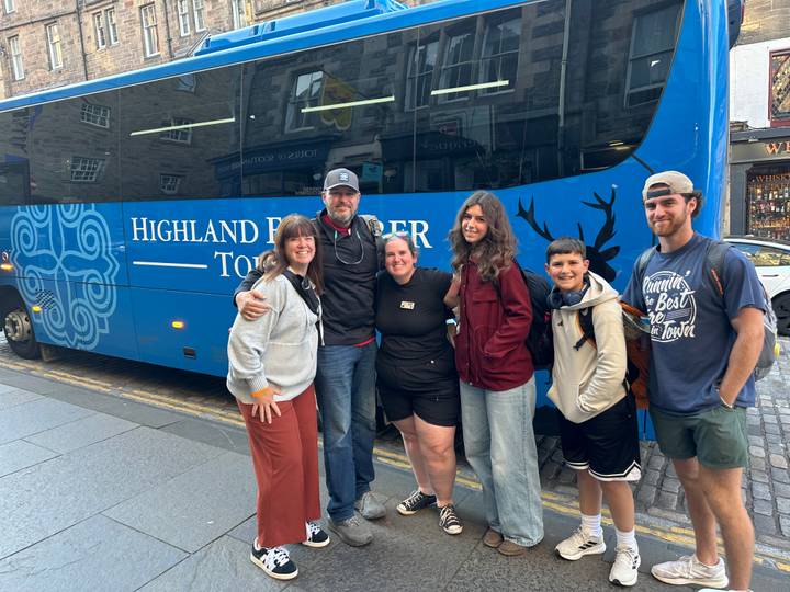 Smiling tour group posing in front of a blue Highland Explorer Tours coach on a city street.