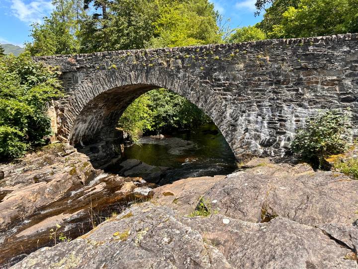 Old stone arch bridge spanning a shallow rocky stream in sunny woodland.