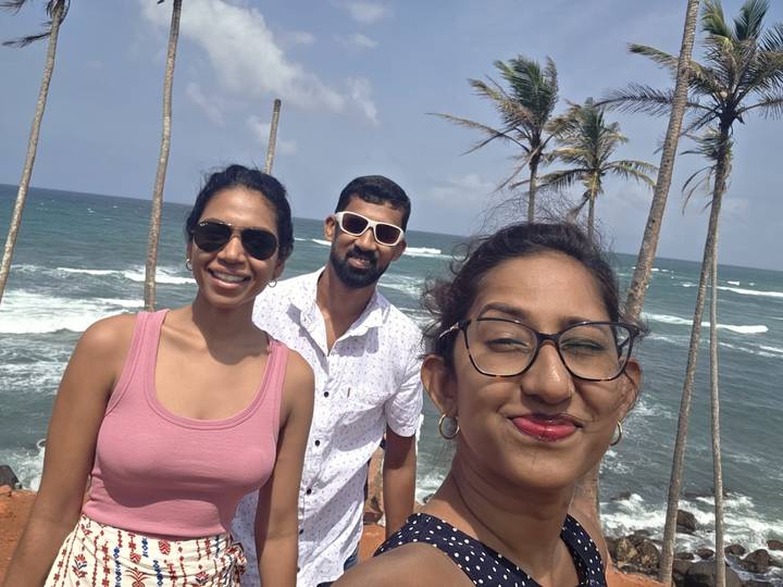 Three friends smile for a selfie on a palm-lined cliff above crashing blue waves on a sunny tropical day.