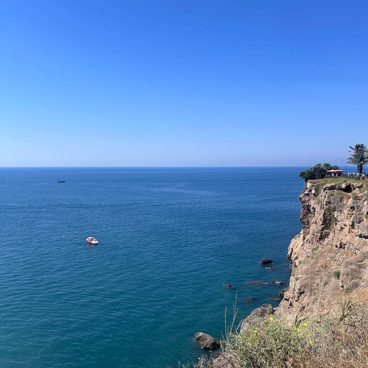 Vast blue Mediterranean stretching to the horizon with a lone boat near a rocky cliff topped by palms.