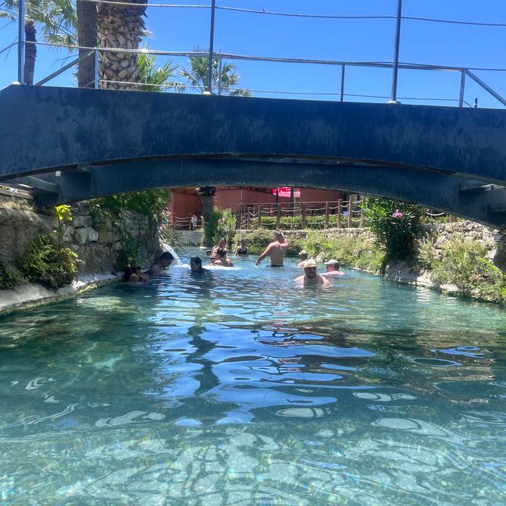 Travellers cool off in a shallow turquoise canal beneath a small bridge on a sunny day.