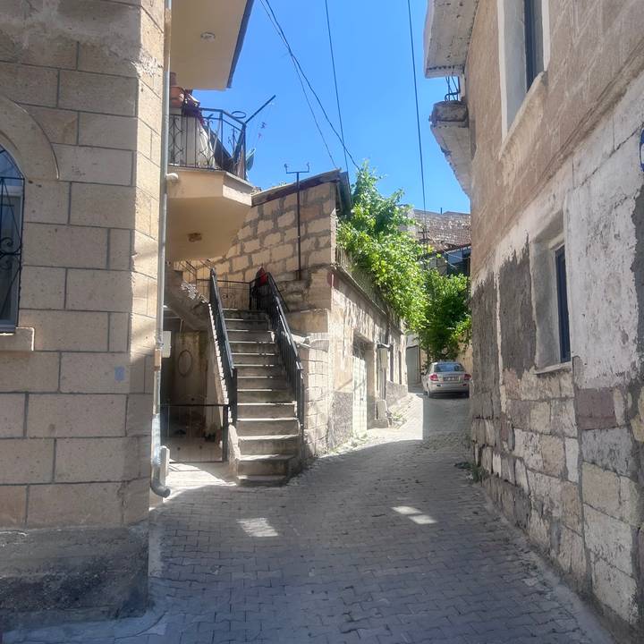 Quiet stone alleyway with external staircase, climbing vines and a parked car in a Cappadocian village.