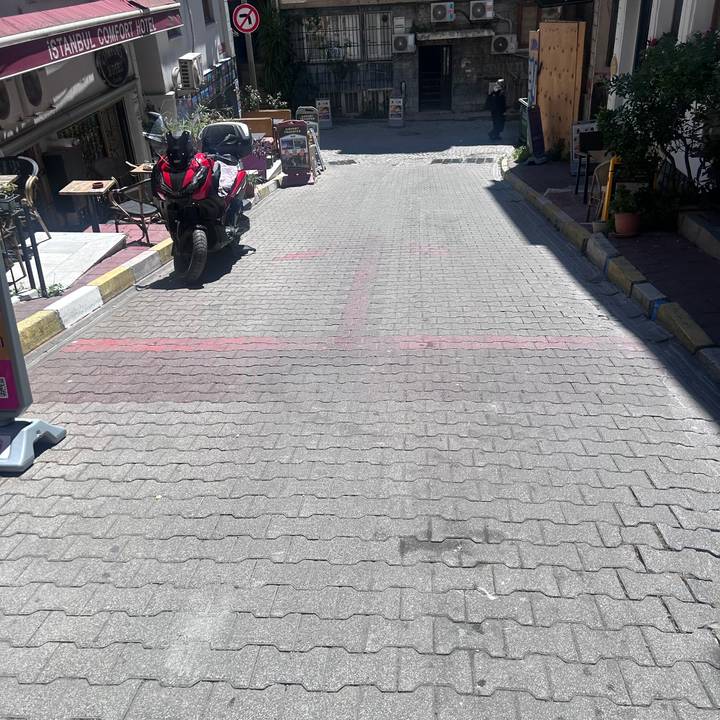 Sun-drenched cobblestone street with red painted arrows, a motorcycle and café terrace tables.