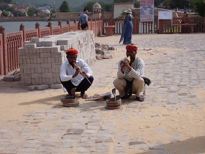 Two snake charmers in colorful turbans perform with flutes and cobra baskets on a sandstone terrace.
