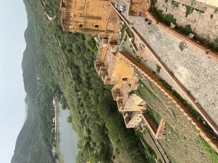 Elevated view of ancient fort walls and yellow sandstone buildings surrounded by green hills and a lake.