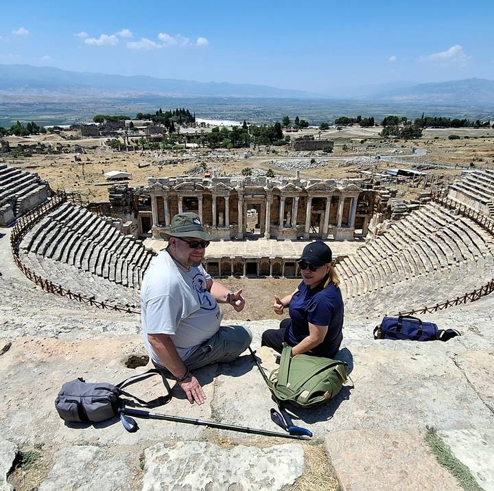 Two tourists sit on the ancient stone tiers of Hierapolis theatre overlooking the Roman stage and surrounding ruins in Pamukkale.