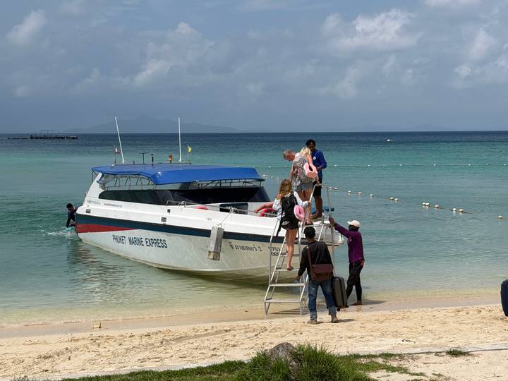 Travellers climb a ladder to board a speedboat anchored off a white-sand beach in clear turquoise water.