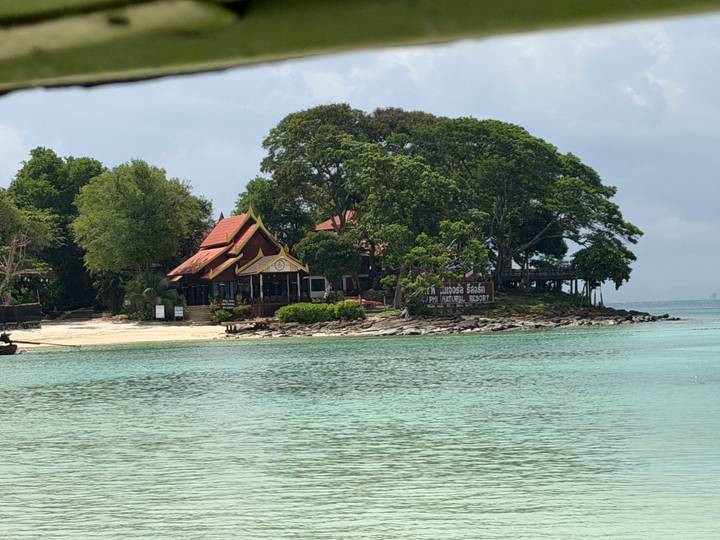 A small tree-covered island with a Thai pavilion sits offshore in calm jade-green sea.