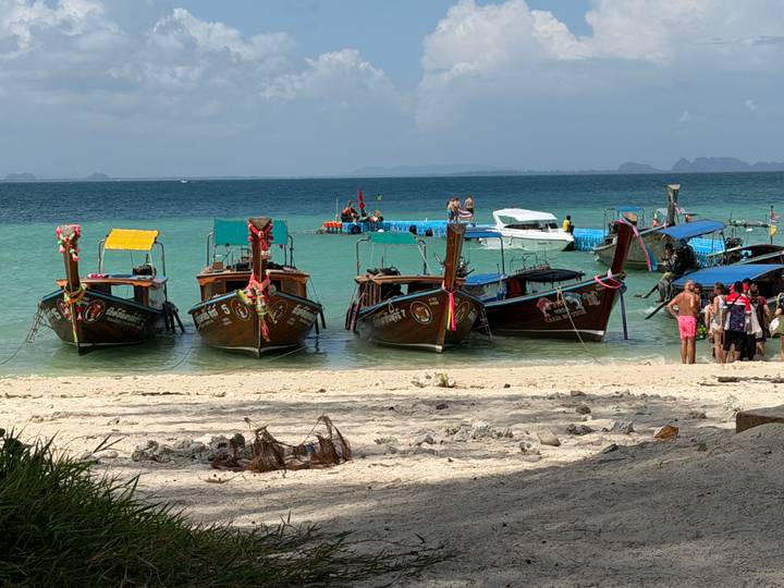 Four colorful long-tail boats tied up along a white-sand beach with turquoise water and distant limestone islands.
