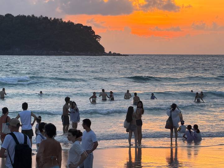 Crowds of swimmers enjoy gentle waves under a vibrant orange sunset sky at a tropical beach.