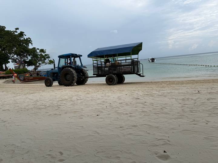 A blue tractor pulling an open passenger cart sits on a wide sandy beach near calm water.