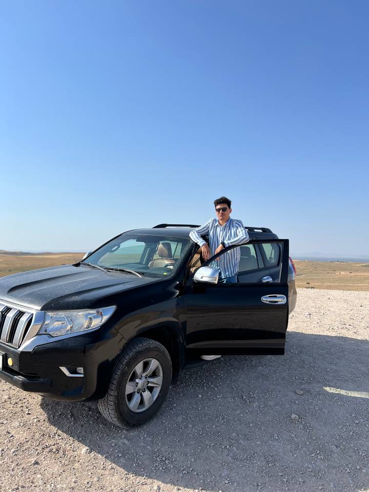Man in sunglasses leans casually on a black 4x4 parked in a wide arid landscape beneath a clear sky.