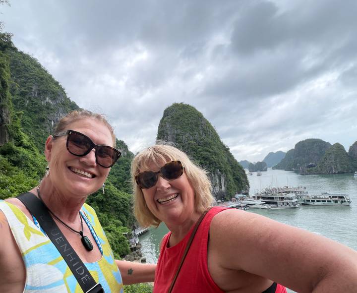 Two smiling women take a selfie with the limestone pillars and tour boats of Halong Bay in the background.
