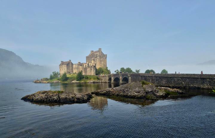 Eilean Donan Castle rises from mist above a stone bridge and reflective tidal waters.