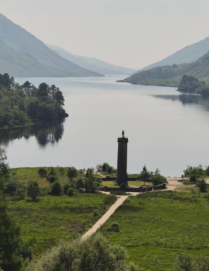 Tall stone Glenfinnan Monument stands beside a tranquil loch surrounded by forested hills.