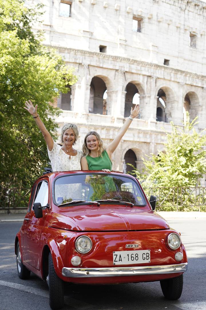 Smiling mother and daughter wave from the roof of a red vintage car parked near an ancient stone amphitheatre.