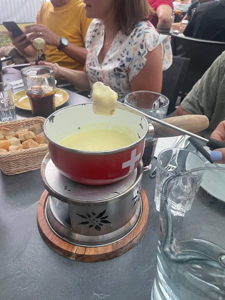 A red fondue pot filled with melted cheese sits on a burner surrounded by cubes of bread and drinks on an outdoor restaurant table.
