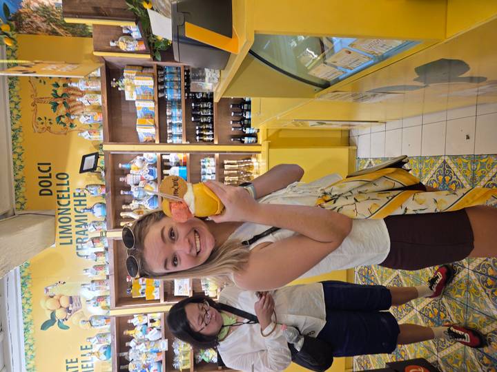 A young woman happily shows a colorful ice-cream and sorbet cup inside a bright yellow limoncello dessert shop.