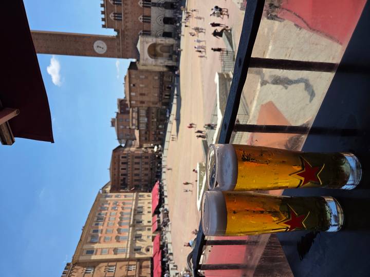 Two tall glasses of beer rest on a balcony railing overlooking a sunny Italian piazza filled with historic brick buildings.