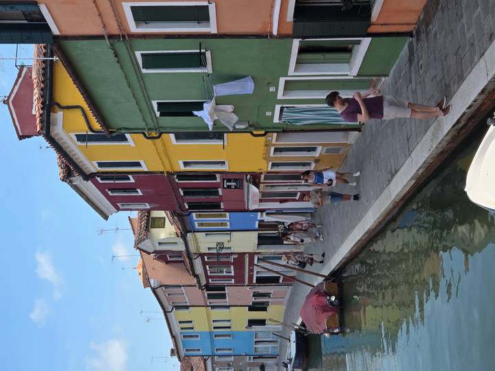 Tourists stroll a canal-side lane lined with brightly painted houses in vivid reds, yellows and greens.