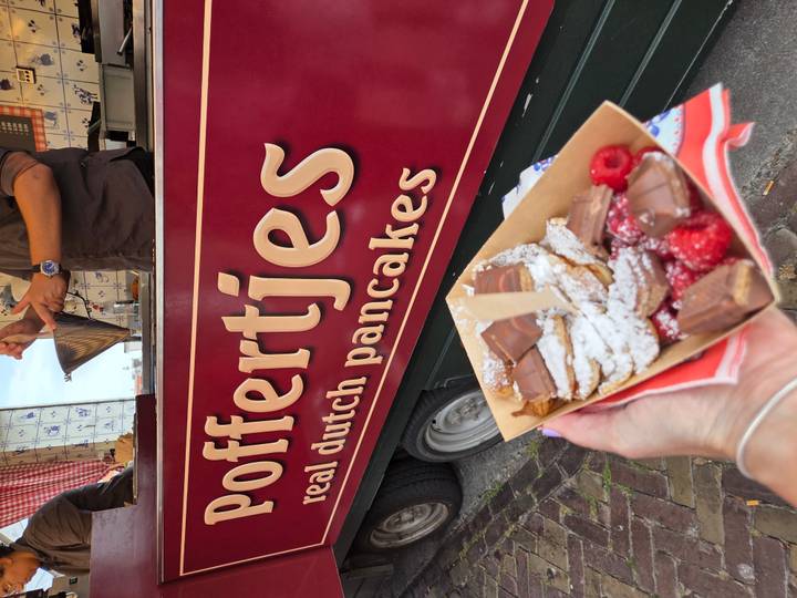 A tray of freshly cooked poffertjes dusted with powdered sugar is held up in front of a maroon Dutch pancake food truck.