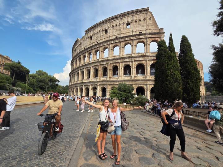Crowds gather outside the Colosseum while two cheerful women pose in front pointing toward the ancient arena.