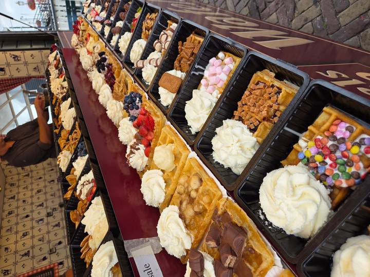 Rows of decadent Belgian waffles topped with whipped cream, fruit and candy fill a market counter display.
