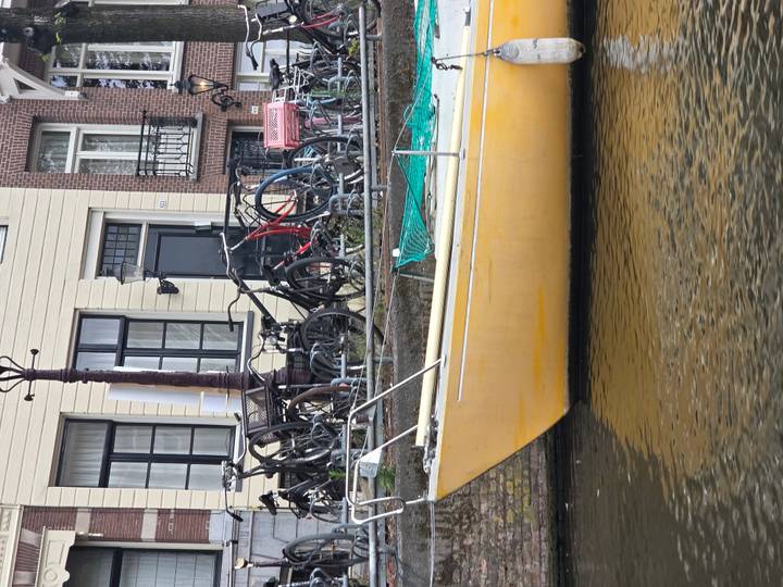 A yellow sailboat is moored along an Amsterdam canal with bicycles lined up on the quay and classic brick houses behind.