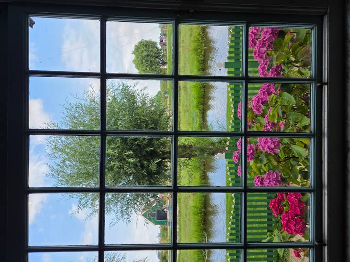 Through a pane-grid window, pink hydrangeas and a willow tree frame a quiet Dutch canal and green pasture.
