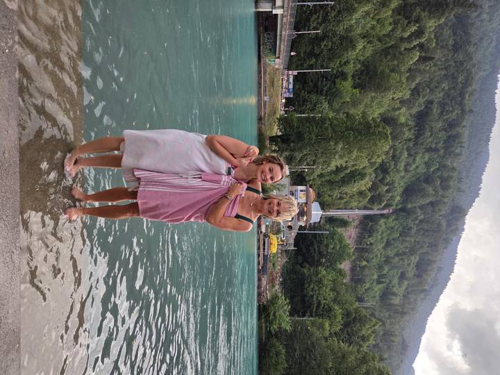Two women wrapped in towels stand ankle-deep in a turquoise alpine river with forested mountains rising behind.