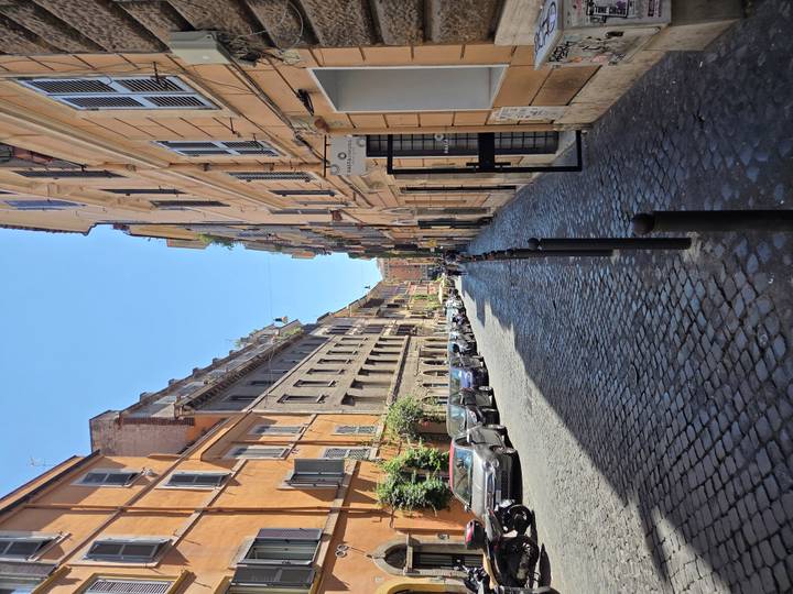 A long, narrow cobblestone street lined with colorful Roman apartment buildings stretches into the distance under a blue sky.