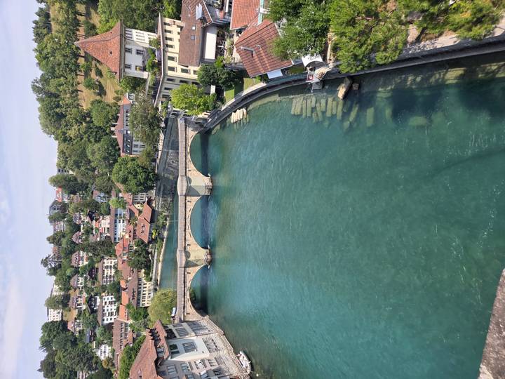 A clear turquoise river flows beneath a stone bridge toward a hillside town of chalet-style homes and trees.