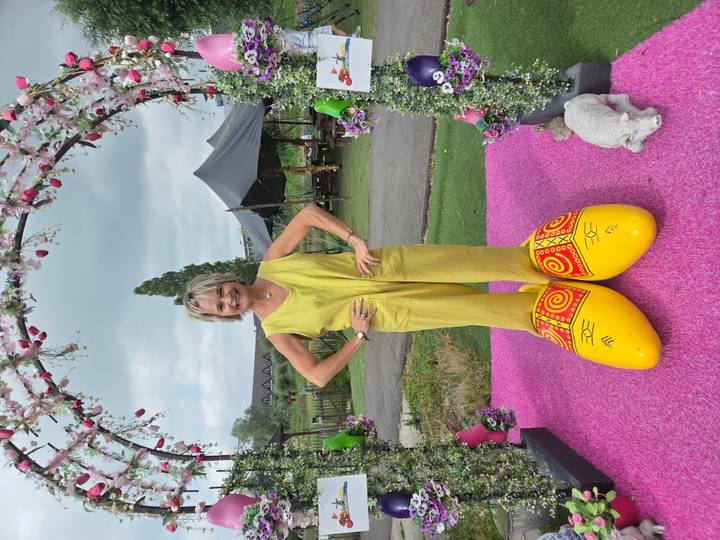 A woman stands on bright pink carpet wearing enormous decorative yellow wooden clogs under a floral arch.