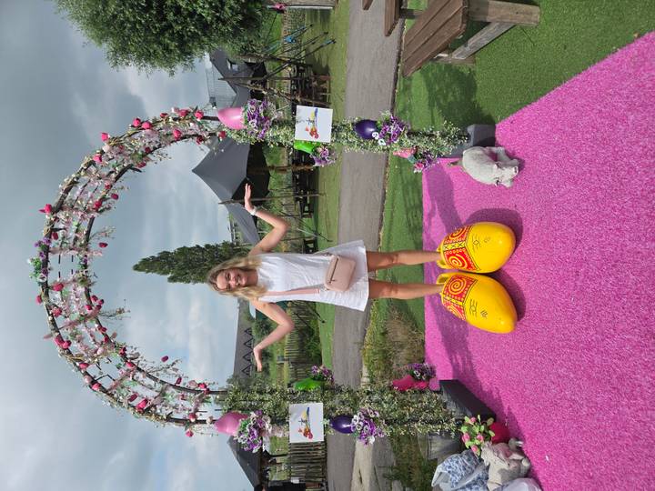 A young woman in a white dress balances inside oversized wooden clogs on a pink mat beneath a flower arch.